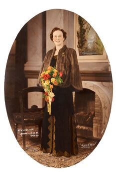 Oval shaped full length studio portrait depicting a standing  woman in a dark evening dress and fur cape. the woman is holding and pearls holding a bouquet of mixed coloured flowers. The woman is standing in front of a fireplace next to a chair.