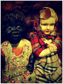 A photograph features two vintage dolls sitting side by side against a dark, textured background. The doll on the left has dark, glossy skin, curly black hair adorned with a red mesh accessory, and an open-mouthed expression. It wears a green floral dress with pink buttons and ruffled details. The doll on the right has a pale yellowish complexion with a cracked and partially broken head, exposing the material beneath. It is wrapped in a red and yellow plaid fabric, covering most of its body. The dolls appear to be posed closely together, conveying an eerie and thought-provoking composition.