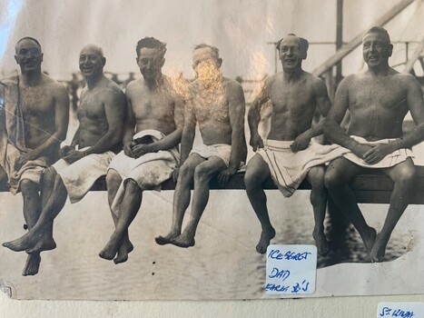 Black and white photograph of six men wearing only towels sitting on what appears to be a pier, with their legs dangling over the water.