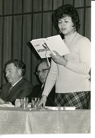 A woman in her 30s reads from a book while two men sit next to her.