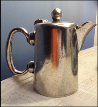 A silver plate milk jug, with a handle, and spout.  A lid with a round knob for opening is attached.