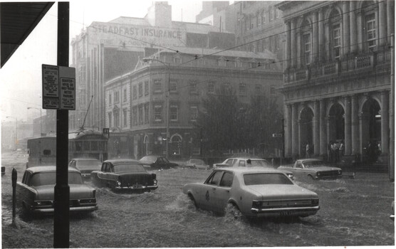 A flooded city street with cars driving through deep water under heavy rain, with buildings and a tram in the background.