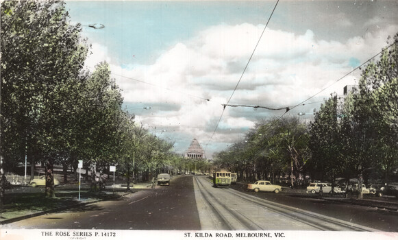 A vintage postcard view looking down St Kilda Road in Melbourne, featuring a tram, cars, and a grand building in the distance framed by trees and overhead tram wires.