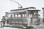 Vintage electric tramcar with open driver's cabin and a trolley pole, parked on tracks next to a brick pillar, with two individuals standing near the tram's front and another near the driver.