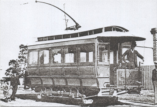 Vintage electric tramcar with open driver's cabin and a trolley pole, parked on tracks next to a brick pillar, with two individuals standing near the tram's front and another near the driver.