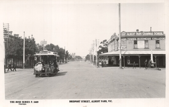 An early 20th-century street scene with a cable tram on Bridport Street, Albert Park, Victoria, featuring buildings on either side of the road including "Albert Park Grammar School" and "Toms' Business College" on the right, and a "J. BOGAN CHEMIST" sign on the left.
