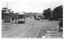 Photograph of a tram turning a corner on St Kilda Road, from The Junction, St Kilda, Victoria. A tram shelter with signage is to the left, with two figures in uniform standing beside it and the tram. The road is wet and reflective. Several early automobiles and a horse-drawn carriage are visible in the distance, navigating the street lined with trees and buildings.