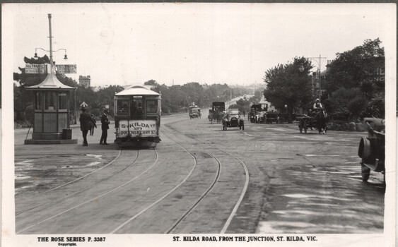 Photograph of a tram turning a corner on St Kilda Road, from The Junction, St Kilda, Victoria. A tram shelter with signage is to the left, with two figures in uniform standing beside it and the tram. The road is wet and reflective. Several early automobiles and a horse-drawn carriage are visible in the distance, navigating the street lined with trees and buildings.