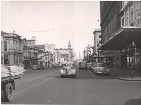 A busy city street with vintage cars and buildings. A truck loaded with timber is on the left side of the street.