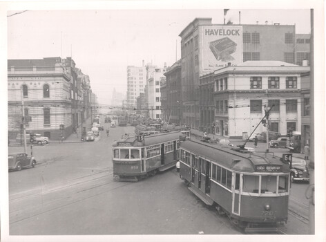 A bustling city street scene with trams and cars. A large advertisement for "HAVELOCK" tobacco is visible on the side of a building.