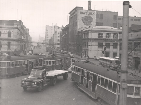 A busy city street with multiple trams, a truck with a trailer, and a bus. The scene is bustling with activity, suggesting a lively urban environment.