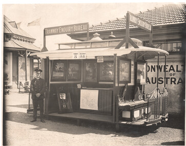 A man in uniform stands beside a tram-shaped booth labeled "Tramway Enquiry Bureau," displaying maps and framed pictures of scenery, with a large sign reading "COMMONWEALTH OF AUSTRALIA" in the background.