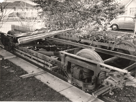 Close-up black and white photo of a disassembled tram bogie with large wheels and springs, resting on wooden planks on the ground.