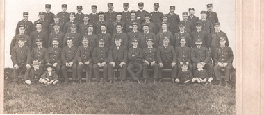 A group of men in uniform are seated on benches outdoors, with grass to the right.