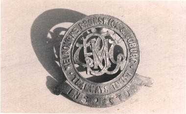 A circular metallic badge with ornate letters MBCTT in the center, surrounded by the text "MELBOURNE BRUNSWICK & COBURG TRAMWAY TRUST INSPECTOR" in a circular pattern.