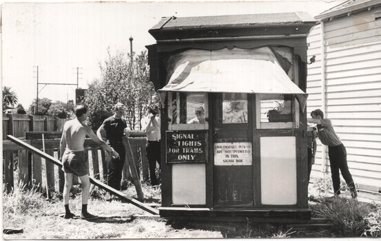 A group of young men stand around a wooden signal box with signs indicating "SIGNAL LIGHTS FOR TRAMS ONLY" and "UNAUTHORISED PERSONS ARE NOT PERMITTED IN THIS SIGNAL BOX."