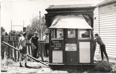 A group of young men stand around a wooden signal box with signs indicating "SIGNAL LIGHTS FOR TRAMS ONLY" and "UNAUTHORISED PERSONS ARE NOT PERMITTED IN THIS SIGNAL BOX."