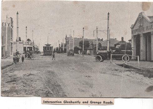 A black and white photograph shows a bustling street scene at an intersection, with a tram, a fire truck, and several early automobiles visible, along with buildings, including a fire brigade station.
