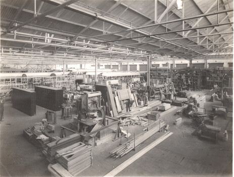 A wide shot of a busy factory or workshop interior with rows of unfinished vehicle or tramcar bodies on the left, and workers and woodworking equipment filling the center and right of the frame.