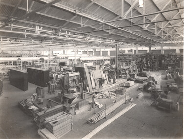 A wide shot of a busy factory or workshop interior with rows of unfinished vehicle or tramcar bodies on the left, and workers and woodworking equipment filling the center and right of the frame.