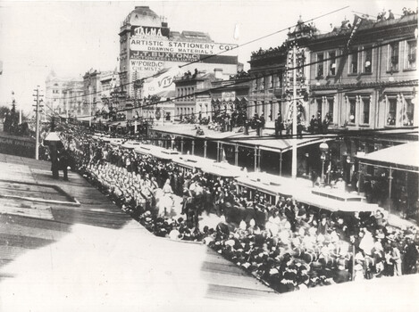 A long procession of trams filled with passengers moves down a street lined with onlookers and buildings. 