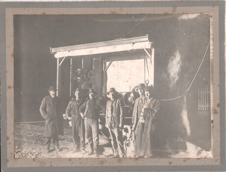 A group of six men in hats and work clothes stand in front of a large machine on tram tracks. 