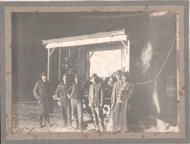 A group of six men in hats and work clothes stand in front of a large machine on tram tracks. 