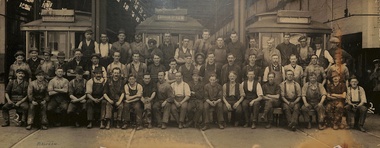 A large group of men in work clothes and hats pose for a photograph in front of three tram cars inside a workshop.