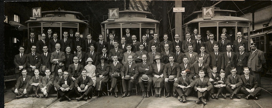 A large group of men, dressed in suits and ties, are posed for a photograph inside and around three different tramcars.