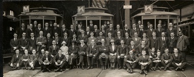 A large group of men, dressed in suits and ties, are posed for a photograph inside and around three different tramcars.