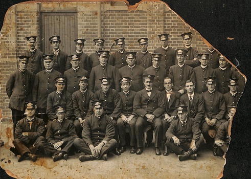 A vintage, worn photograph depicts a formal group portrait of many young men in matching dark uniforms with peaked caps, posing in front of a brick building.