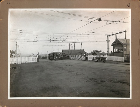 A street scene with a railway signal box and a white picket fence bordering several buildings, including a tramcar, shed and houses. Overhead electrical wires for an electric railway and tramway system crisscross the sky above the street. A vintage car is parked near the fence.