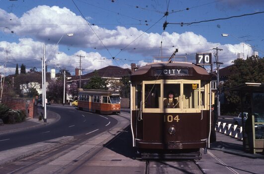 Two brown and orange trams are on a city street with overhead wires. The tram in the foreground has "CITY" above the windshield and the number "104" on its front. The tram in the background is further down the street, rounding a curve. Houses and trees line the street. The sky is blue with fluffy white clouds.