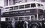 A vintage double-decker bus with "MELBOURNE & METROPOLITAN TRAMWAYS BOARD." written in white on its side, stands in front of a brick building. Four men in formal attire stand beside the bus.