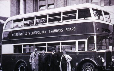 A vintage double-decker bus with "MELBOURNE & METROPOLITAN TRAMWAYS BOARD." written in white on its side, stands in front of a brick building. Four men in formal attire stand beside the bus.