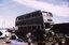 A derelict green and white double-decker bus sits in a junkyard surrounded by scrap metal and car parts under a cloudy sky.