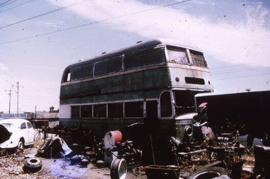 A derelict green and white double-decker bus sits in a junkyard surrounded by scrap metal and car parts under a cloudy sky.