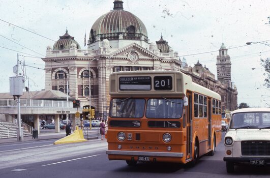 An orange bus with the route number 201 displayed on its front drives past a grand building - Flinders St station  - with a large dome. In the foreground, part of a white van is visible.
