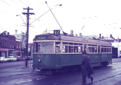 A green tram numbered 53 and route 82 stops on a wet street in front of shops while a man in a trench coat walks away from the tram. A billboard above the tram reads "Sentence it to 4 years hard labour in your car."