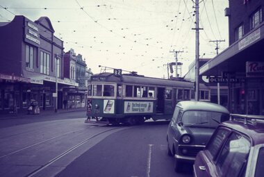 A vintage green tram with advertisements travels down a city street lined with buildings. Cars and pedestrians are visible on the street as overhead wires crisscross the sky.