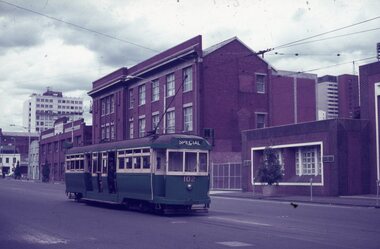 A green and cream vintage tram numbered 102 labeled "SPECIAL" travels down a city street lined with brick buildings and a modern high-rise in the background under a cloudy sky.