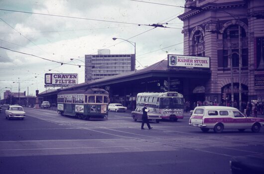 A green tram and various vehicles are seen at a city intersection, with large billboards advertising "CRAVEN FILTER" and "KAISER STUHL COLD DUCK" overhead. A police man walks across the street in front of a bus and a white utility vehicle.