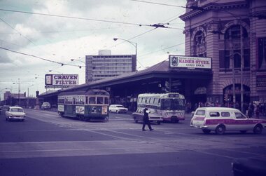 A green tram and various vehicles are seen at a city intersection, with large billboards advertising "CRAVEN FILTER" and "KAISER STUHL COLD DUCK" overhead. A police man walks across the street in front of a bus and a white utility vehicle.