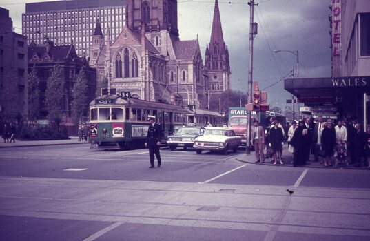 A policeman directs traffic at a busy intersection in front of a cathedral, with a tram and cars visible. People gather on the footpath near a building labeled "WALES".
