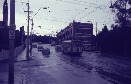 A vintage green tram travels down a wet, grey street lined with cars and buildings under an overcast sky, with a sign on a pole reading "HAIL CARS HERE".