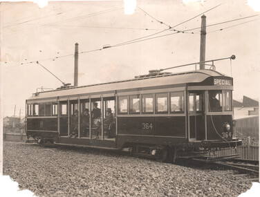 A vintage electric tram car, numbered 364 and labeled "SPECIAL," sites on tracks surrounded by gravel. Several passengers, dressed in period attire, are seated inside the open-sided tram. The tram is connected to overhead wires by a trolley pole. 