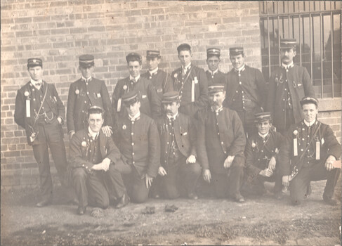 A group of fourteen men in uniform, some standing and some kneeling, pose for a sepia-toned photograph in front of a brick wall. The men are wearing dark jackets, high-necked shirts with ties, and peaked caps. Many of the jackets are adorned with decorative ribbons and medals. The group appears to be a formal organization or team.