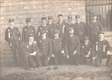 A group of fourteen men in uniform, some standing and some kneeling, pose for a sepia-toned photograph in front of a brick wall. The men are wearing dark jackets, high-necked shirts with ties, and peaked caps. Many of the jackets are adorned with decorative ribbons and medals. The group appears to be a formal organization or team.