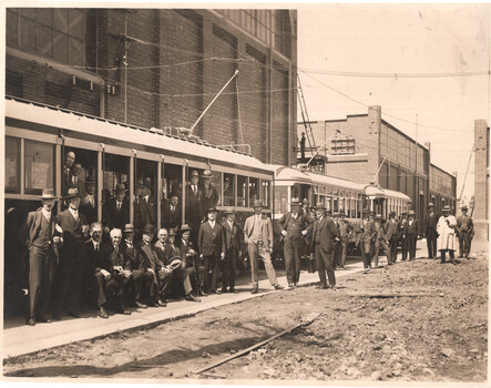 A group of men in suits and hats stand alongside two tramcars in front of a brick building.