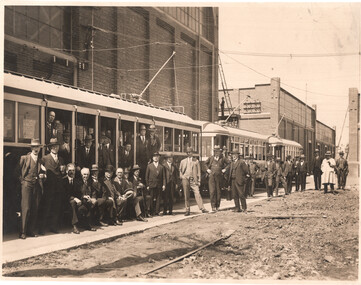 A group of men in suits and hats stand alongside two tramcars in front of a brick building.